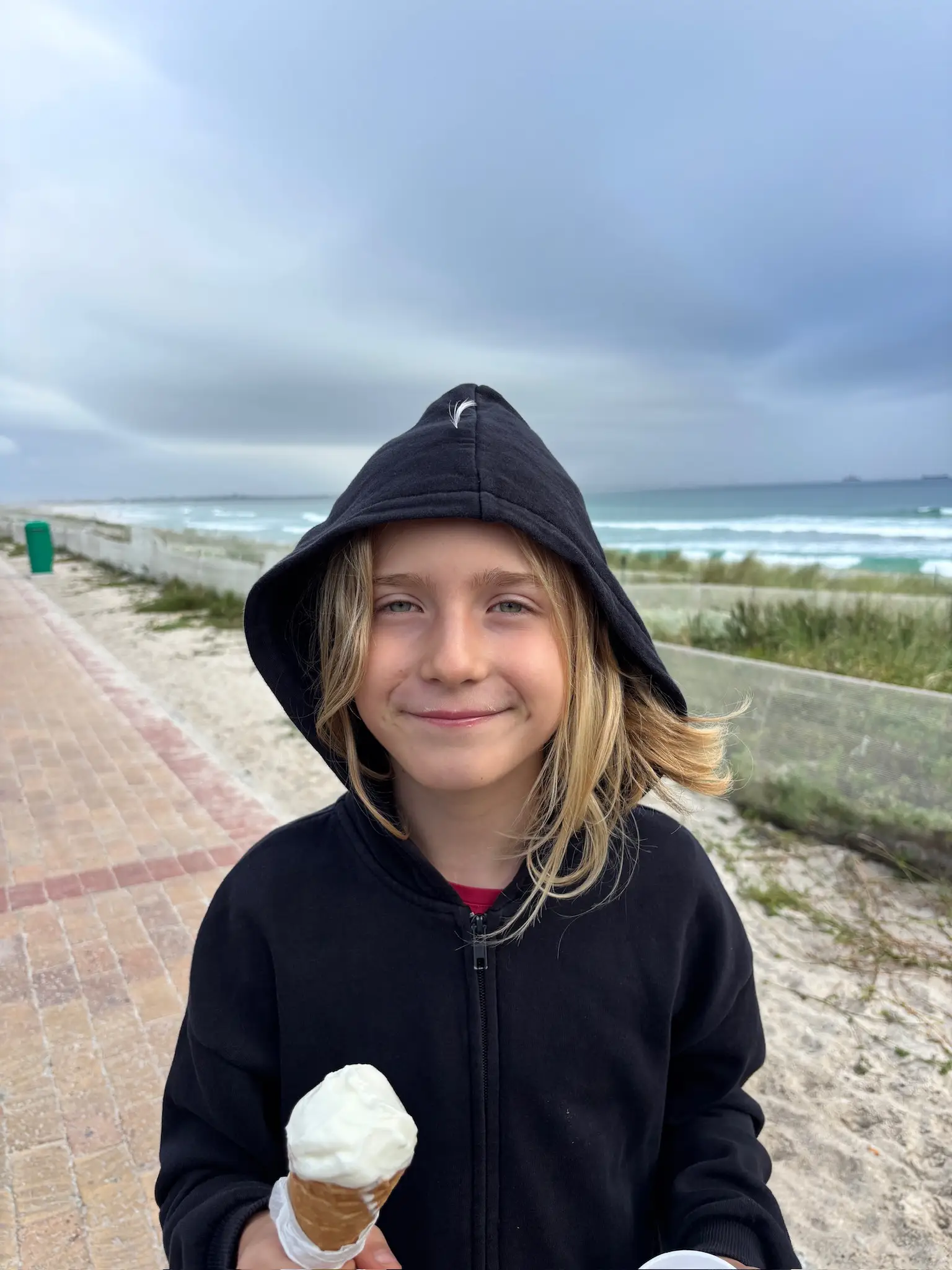 Merle isst Eis in einer dicken Kapuzenjacke am Strand